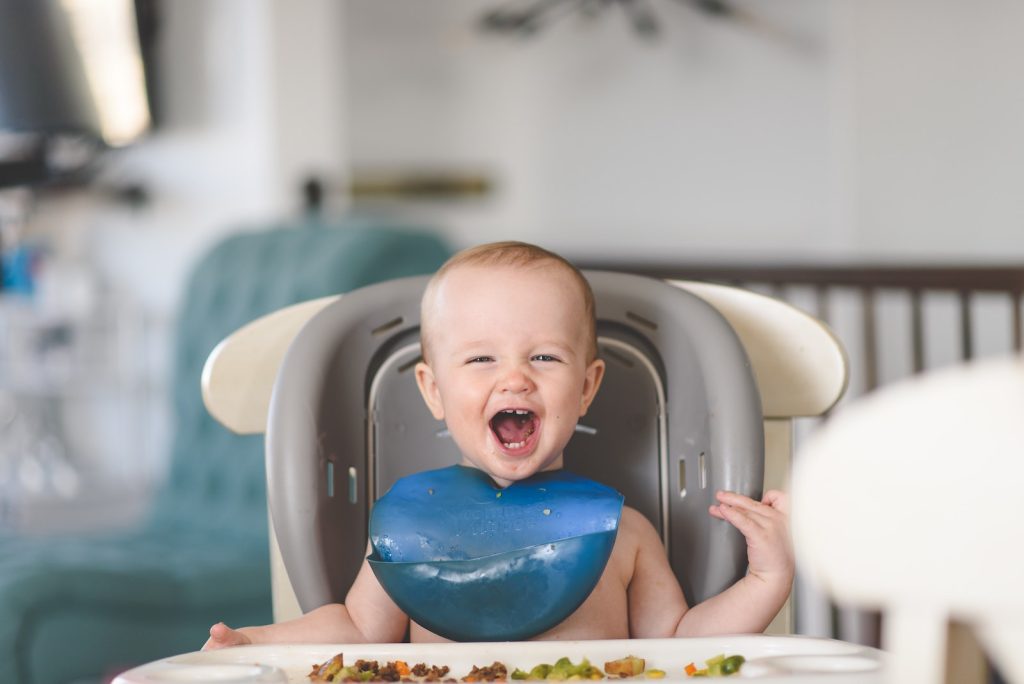 Baby with blue bib on sitting in a gray and white high chair.
